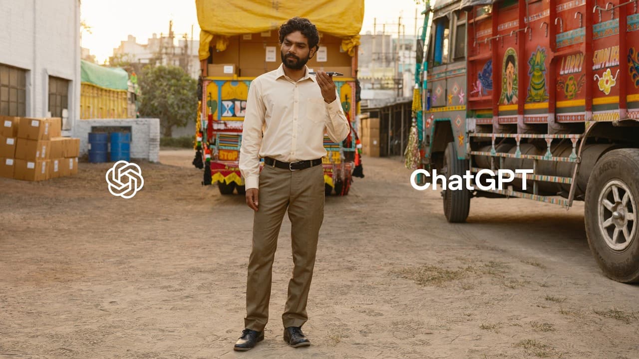 Man in a factory office looking at a smartphone, with boxes and trucks visible in the background.